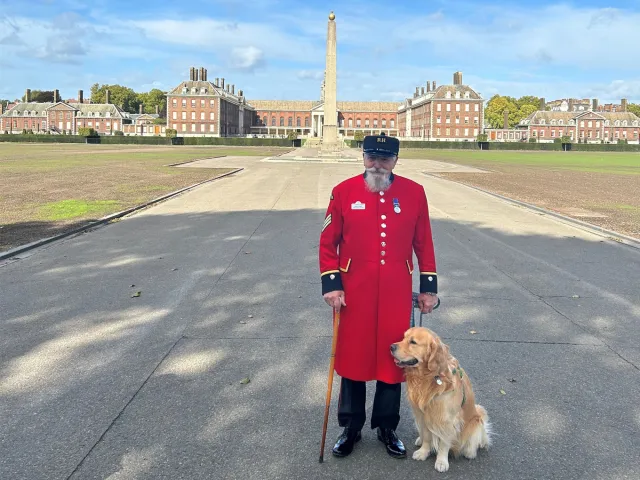 Chelsea Pensioner stands with a golden retriever at Chelsea Dog Day, welcoming visitors.