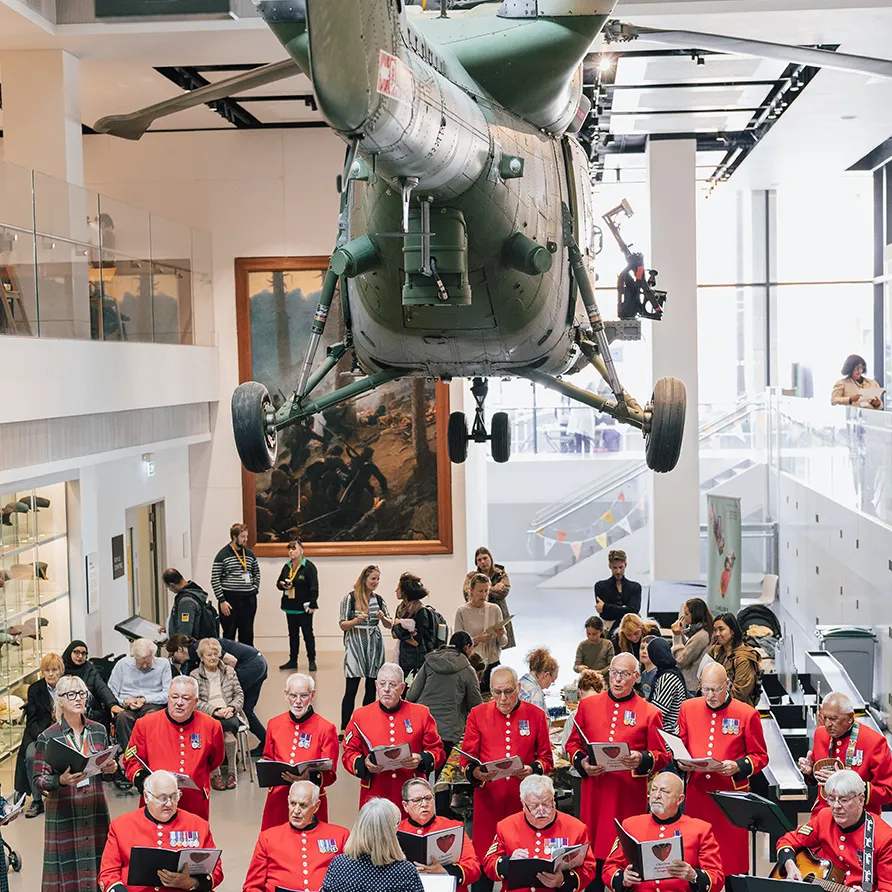 Veteran choir performing under a suspended helicopter in a museum during the Chelsea History Festival.