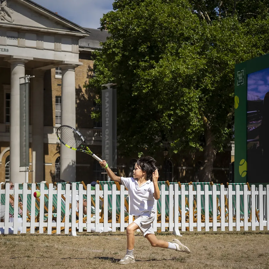 Young child swinging a tennis racket to hit a ball during an outdoor family event.