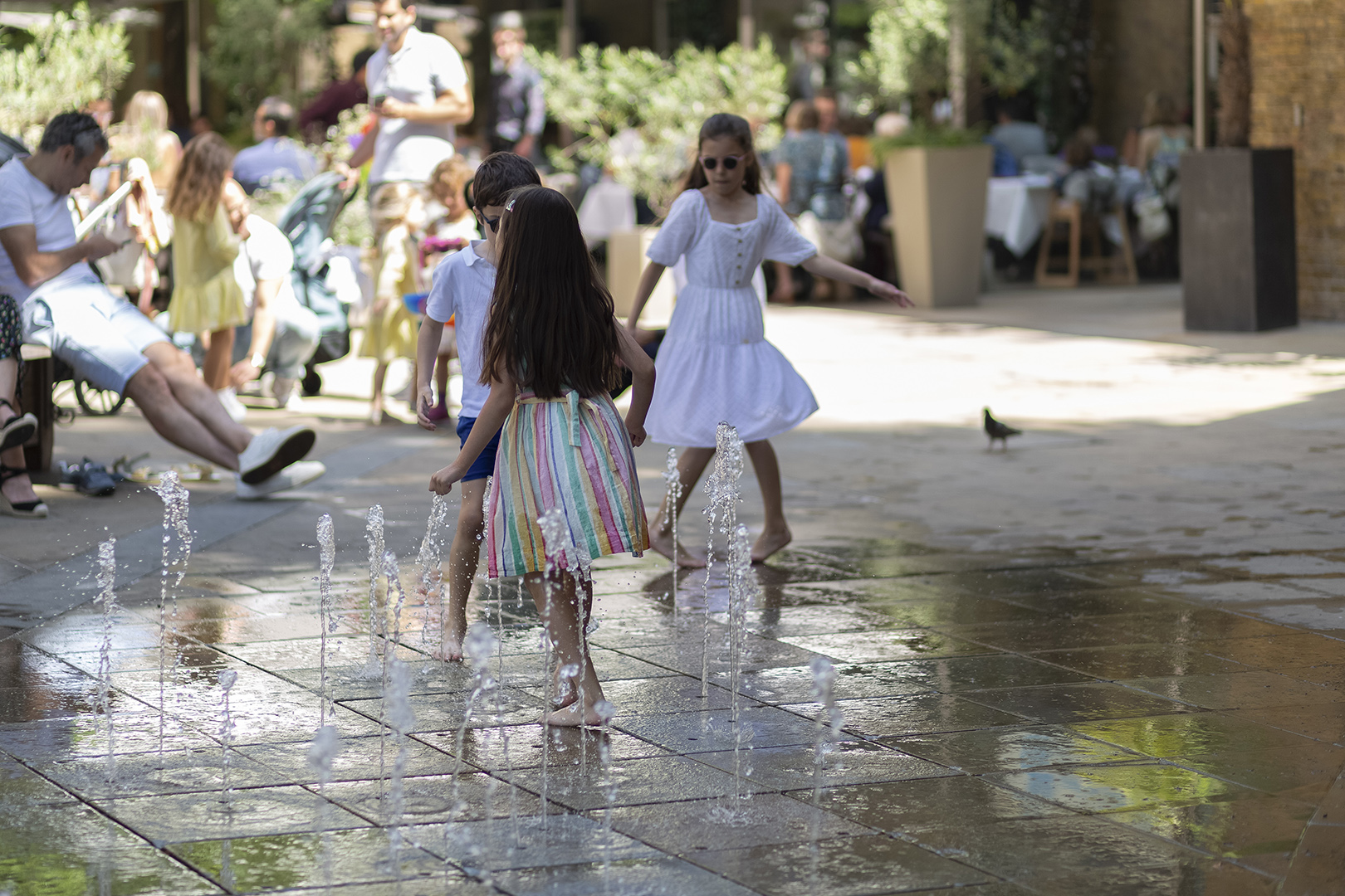 Family enjoying a day out with children playing in a public splash fountain while adults relax nearby.