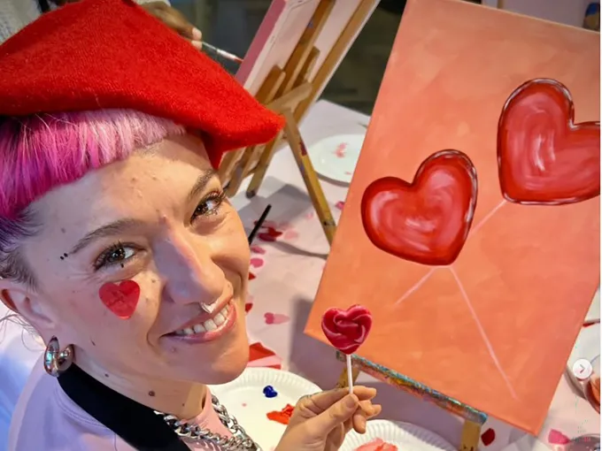 Woman smiling while painting heart-themed canvases at a social painting workshop.