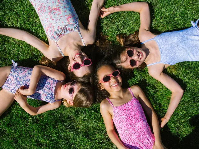 Four children lying on grass in swimsuits and sunglasses enjoying a sunny summer day.